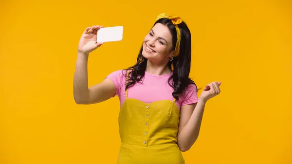 Femme souriante en bandeau prenant selfie sur smartphone isolé sur jaune — Photo de stock