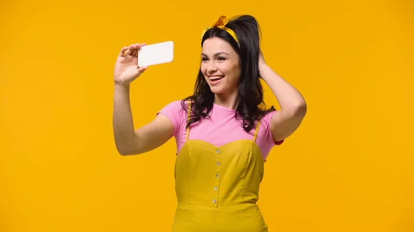 Красивая женщина в повязке на голове регулирует волосы, делая селфи на смартфоне, изолированном на желтый — стоковое фото