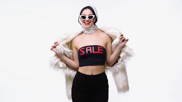 Fröhliche Frau in Sonnenbrille und Top mit Verkaufsaufdruck hält flauschige Jacke isoliert auf weiß — Stockfoto