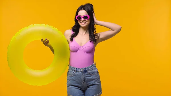 Красивая женщина в солнечных очках и купальнике улыбается, держа кольцо для купания изолированным на желтый — стоковое фото