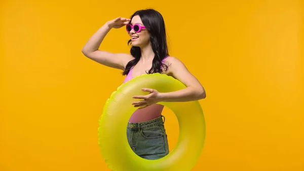 Bruna donna in occhiali da sole tenendo anello di nuoto e guardando lontano isolato sul giallo — Foto stock