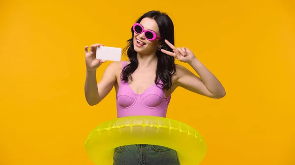 Giovane donna in occhiali da sole e anello di nuoto prendendo selfie e mostrando segno di pace isolato su giallo — Foto stock