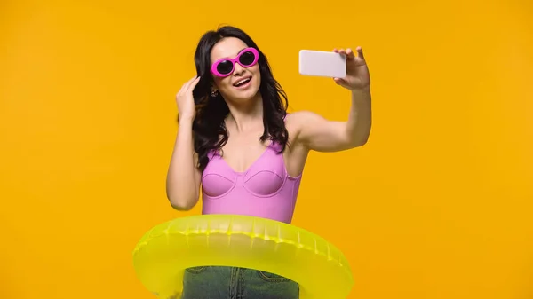 Красива жінка в сонцезахисних окулярах і плавати кільце приймає селфі ізольовано на жовтому — стокове фото