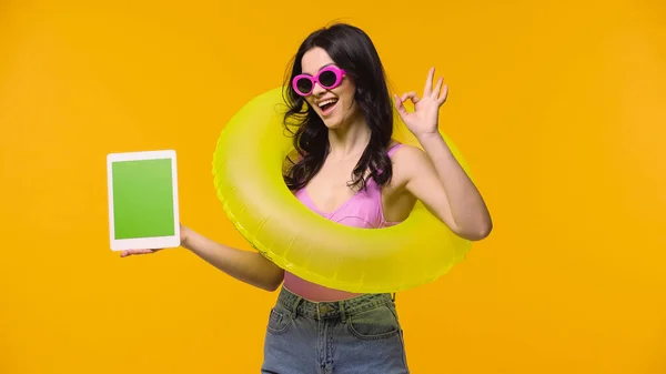 Frau mit Sonnenbrille und Schwimmring zeigt ok, während sie digitales Tablet mit grünem Bildschirm auf gelb isoliert hält — Stockfoto