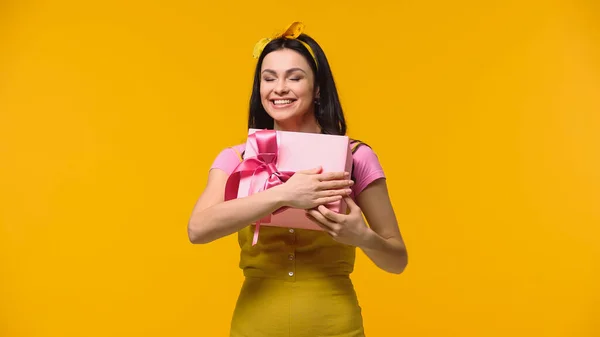Усміхнена жінка з закритими очима тримає подарунок з бантом ізольовано на жовтому — стокове фото