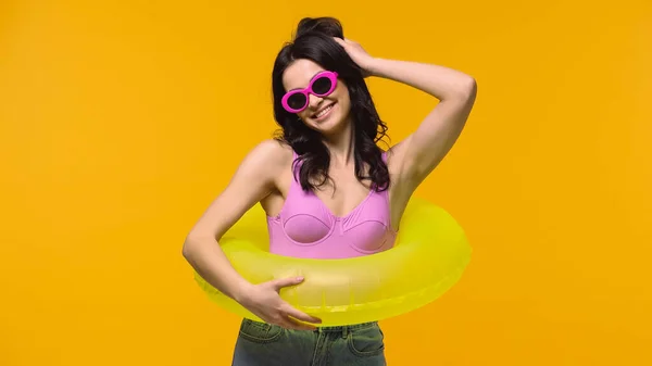 Mujer sonriente en gafas de sol y anillo de natación ajustando el cabello aislado en amarillo - foto de stock