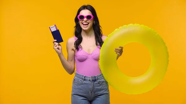 Excited woman in sunglasses holding passport with air ticket and swim ring isolated on yellow — Stock Photo