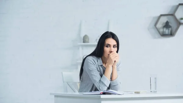 Mujer de negocios preocupada con las manos cerradas sentada en el escritorio en la oficina - foto de stock