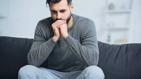 Homem preocupado em camisola cinza sentado com as mãos apertadas no sofá — Fotografia de Stock
