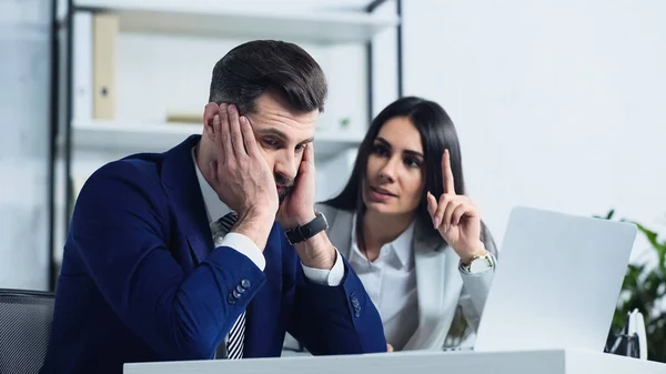 Грустный бизнесмен смотрит в сторону размытой деловой женщины, указывающей пальцем во время ссоры в офисе — стоковое фото