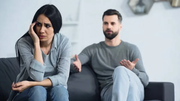 Miserable woman sitting near blurred and displeased boyfriend quarrelling in living room — Stock Photo