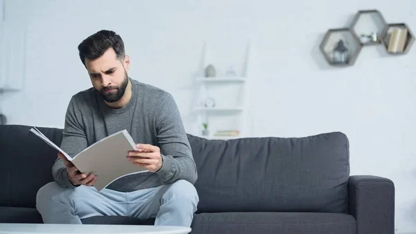 Dissatisfied man looking at folder while sitting on sofa in living room — Stock Photo