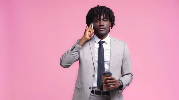 Joven hombre de negocios afroamericano sosteniendo taza de papel y hablando por teléfono celular aislado en rosa - foto de stock
