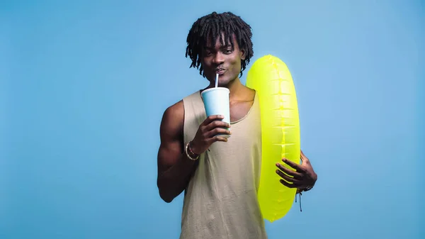 African american man with paper cup drinking soft drink and holding inflatable ring isolated on blue — Stock Photo