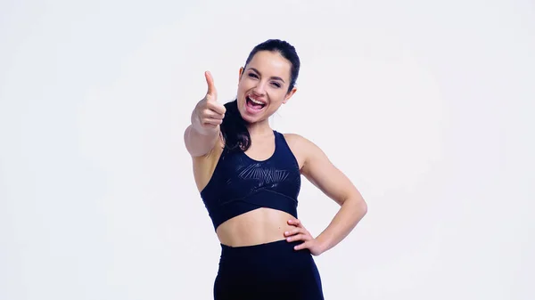 Deportista sonriente mostrando el pulgar hacia arriba mientras está de pie con la mano en la cadera aislado en blanco - foto de stock