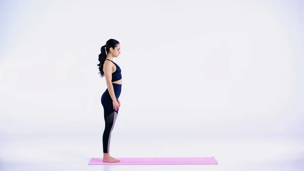 Vista laterale di giovane donna in abbigliamento sportivo in piedi sul tappeto fitness su bianco — Foto stock