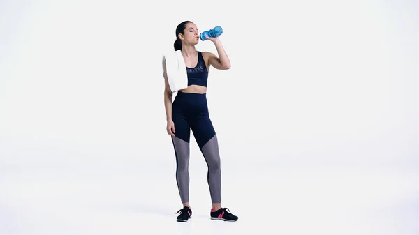 Müde Sportlerin mit Handtuch trinkt Wasser aus Sportflasche auf weiß — Stockfoto