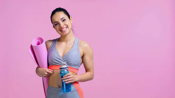 Щаслива спортсменка тримає фітнес-матер і спортивну пляшку ізольовано на рожевому — стокове фото