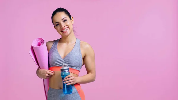 Happy sportswoman holding fitness mat and sports bottle isolated on pink — Stock Photo