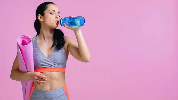 Junge Sportlerin trinkt Wasser aus Sportflasche und hält Fitnessmatte isoliert auf rosa — Stockfoto