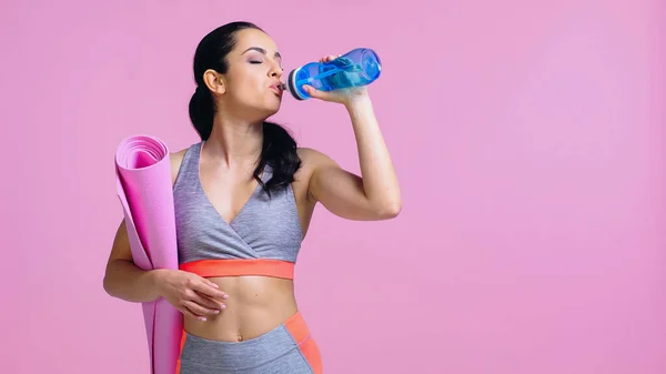 Young sportswoman drinking water from sports bottle and holding fitness mat isolated on pink — Stock Photo