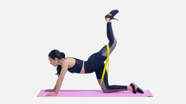 Brunette jeune sportive travaillant avec des élastiques jaunes sur tapis de fitness rose sur blanc — Photo de stock