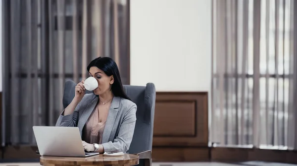 Brunette freelancer using laptop and drinking coffee in lobby of restaurant — Stock Photo