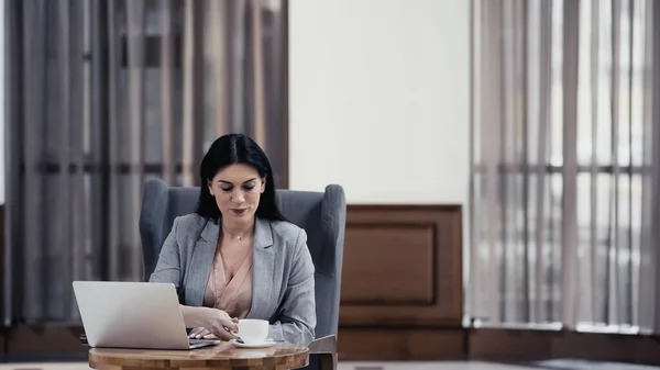 Brunette freelancer reaching cup of coffee near laptop on table in lobby of restaurant - foto de stock