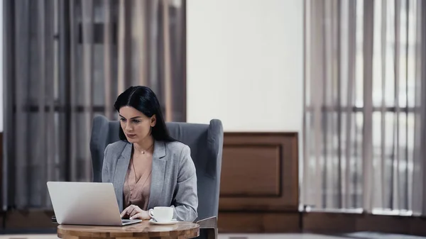 Brunette freelancer typing on laptop near cup of coffee on table in lobby of restaurant — Fotografia de Stock