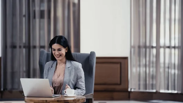 Happy woman looking at laptop near cup of coffee on table in lobby of restaurant — Photo de stock
