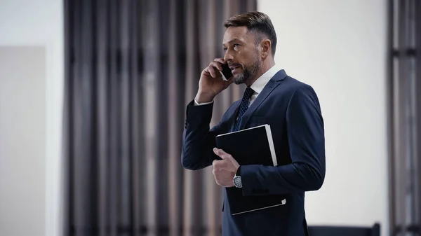 Bearded businessman with paper folder talking on smartphone in lobby of restaurant — Fotografia de Stock