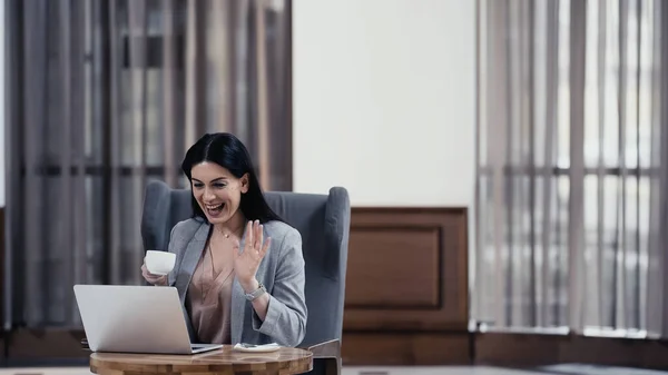 Happy woman holding cup and waving hand during video call on laptop - foto de stock