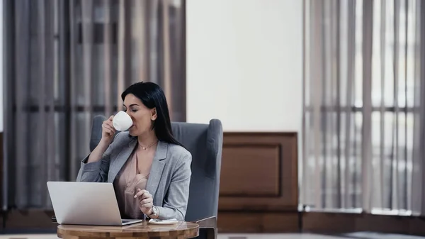 Freelancer drinking coffee near laptop on table in restaurant - foto de stock