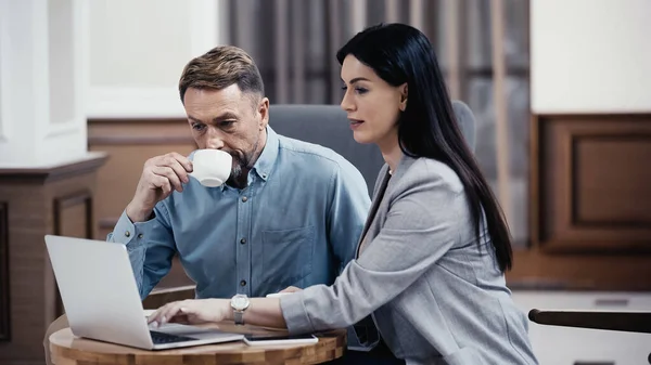 Businessman drinking coffee and looking at laptop near woman in lobby of restaurant — Foto stock