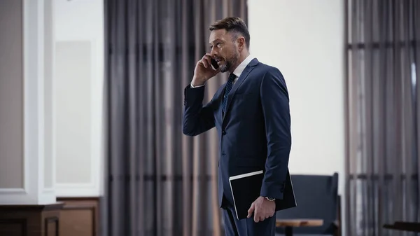 Bearded businessman in suit talking on smartphone and holding paper folder in restaurant — Stock Photo