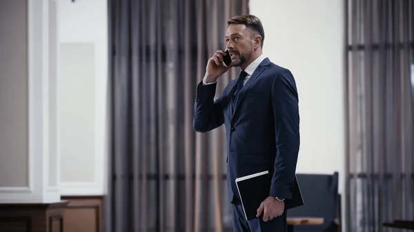 Bearded businessman in suit talking on mobile phone and holding paper folder in restaurant — Stock Photo