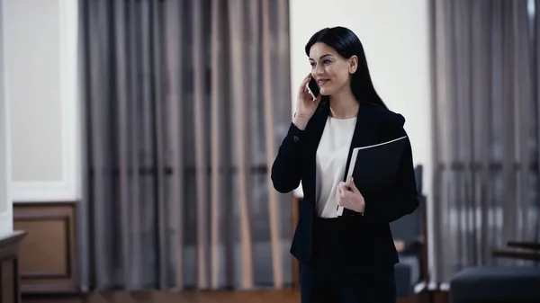 Cheerful businesswoman with paper folder talking on smartphone in lobby of restaurant — Fotografia de Stock