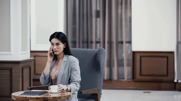 Businesswoman talking on cellphone near cup of coffee in lobby of restaurant — Stock Photo