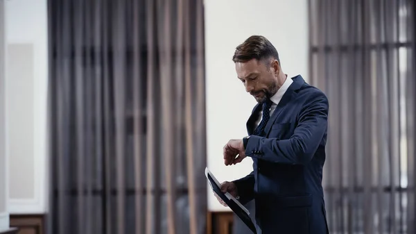 Bärtiger Geschäftsmann schaut auf Uhr und hält Papiermappe in der Hand — Stockfoto