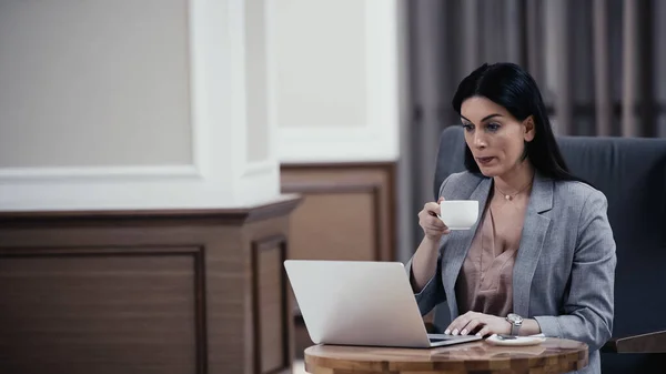Женщина держит чашку, глядя на ноутбук в ресторане — стоковое фото