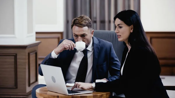 Mann im Anzug trinkt Kaffee in der Nähe von Geschäftsfrau mit Laptop — Stockfoto