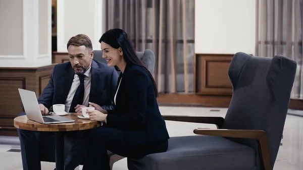 Man in suit and happy businesswoman looking at laptop near cups on table — Fotografia de Stock