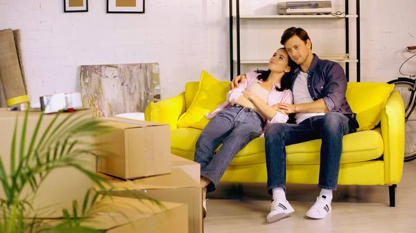 Smiling couple talking and resting on couch near boxes in new home — Stock Photo