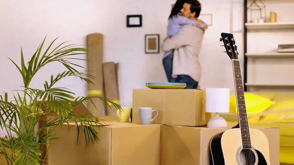 Guitarra acústica, planta y cajas cerca borrosa pareja abrazos en el fondo - foto de stock