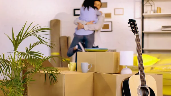 Guitarra acústica, plantas y cajas de cartón cerca de pareja borrosa abrazándose en el fondo - foto de stock
