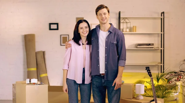 Cheerful man and woman hugging while standing near boxes in new home — Stock Photo