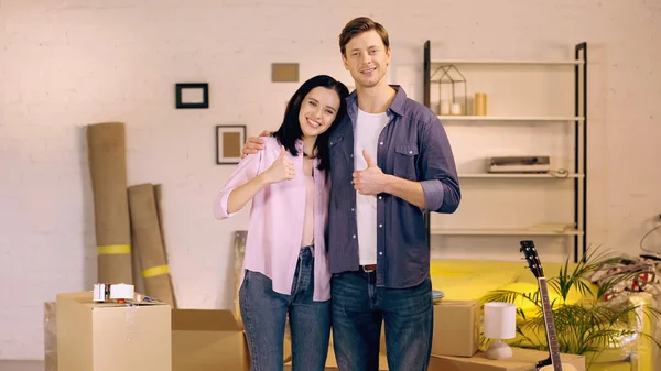 Couple souriant montrant pouces près des boîtes dans la nouvelle maison — Stock Photo