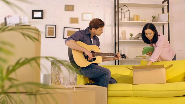 Uomo suonare la chitarra acustica mentre donna disimballaggio libri in nuova casa — Foto stock