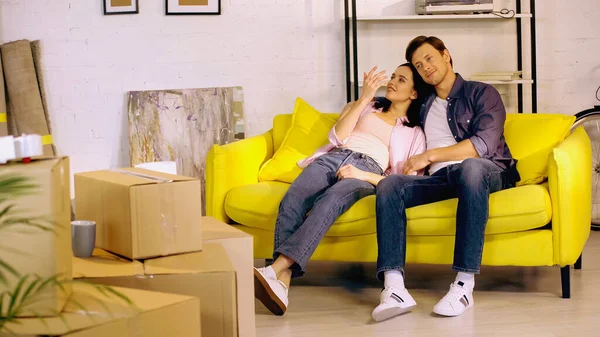 Happy couple talking and resting on couch near boxes in new home — Stock Photo