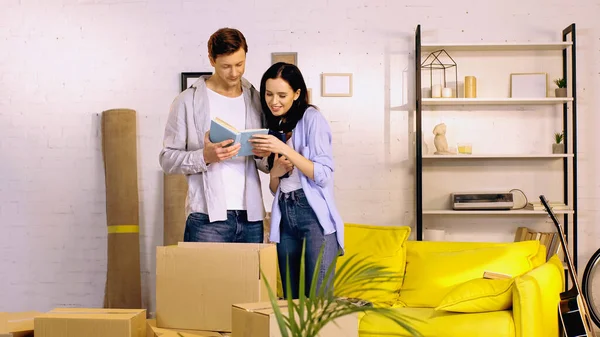 Alegre joven pareja leyendo libro cerca de cajas en sala de estar - foto de stock